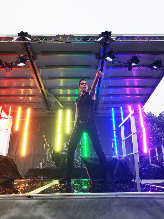 Brie Treviranus striking a pose on stage with the stage lights set to the pride rainbow.