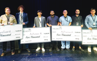 At the Velocity Final Pitch competition, six individuals stand on stage holding large checks from Velocity. Each check is for five thousand dollars, awarded to groups like PyroGuard and Automa AI. They are all smiles against a dark background, celebrating their innovative contributions to firefighter safety.