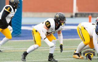 Football players in white and yellow uniforms from the Waterloo Warriors team are lined up on the field, ready for a play. One varsity athlete in a black helmet and jersey number 43 is in a crouched stance. The team's logo and name appear in the bottom right corner, showcasing their UW pride.