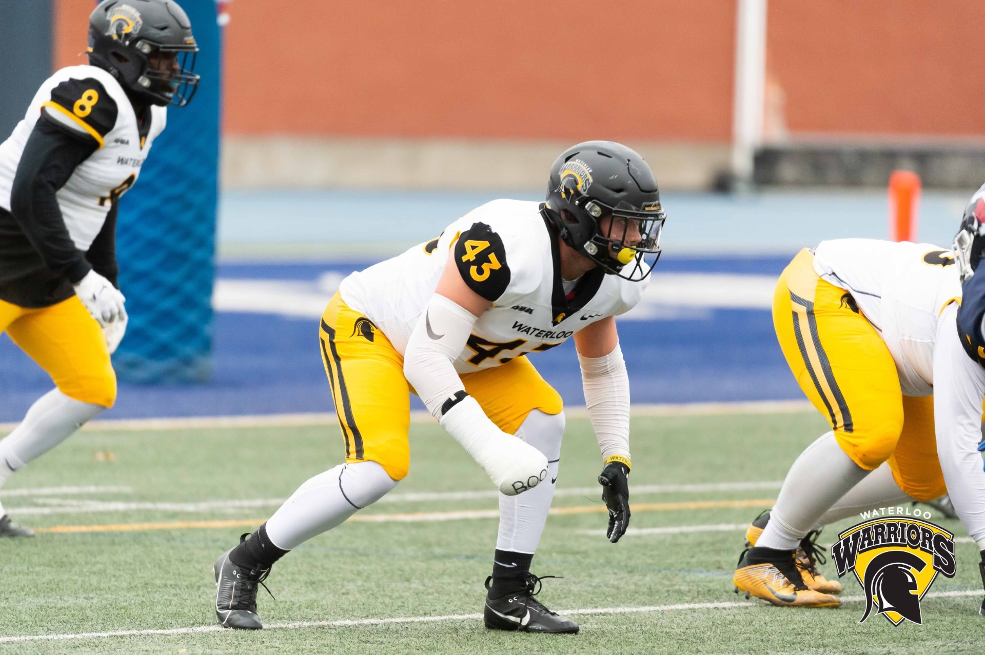 Football players in white and yellow uniforms from the Waterloo Warriors team are lined up on the field, ready for a play. One varsity athlete in a black helmet and jersey number 43 is in a crouched stance. The team's logo and name appear in the bottom right corner, showcasing their UW pride.