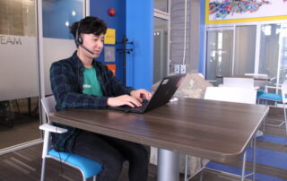 A person wearing a plaid shirt and headset sits at a modern office desk, working on a laptop. The setting includes blue walls, glass doors, and colorful artwork in the background, creating a professional yet vibrant workspace. It's clear they're focused on their next co-op project.