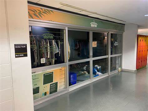 A glass-fronted thrift shop with clothes hanging on racks and various items placed on shelves. The store has a green-themed sign, and a small open sign is visible in one window. There's a black sign on the left wall with "WUSA Thrift" and "SLC 0006" written on it, noting it's closed fall term except pop-up events.