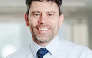 A man with short dark hair and a beard is smiling. He is wearing a light blue dress shirt and a dark blue tie with white dots. The background is blurred, suggesting an indoor setting. This Waterloo researcher was recently awarded $2 million to study the harms of vaping.