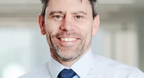 A man with short dark hair and a beard is smiling. He is wearing a light blue dress shirt and a dark blue tie with white dots. The background is blurred, suggesting an indoor setting. This Waterloo researcher was recently awarded $2 million to study the harms of vaping.