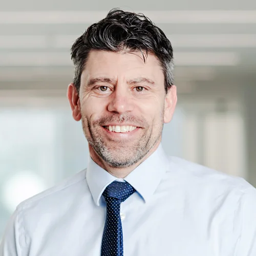 A man with short dark hair and a beard is smiling. He is wearing a light blue dress shirt and a dark blue tie with white dots. The background is blurred, suggesting an indoor setting. This Waterloo researcher was recently awarded $2 million to study the harms of vaping.