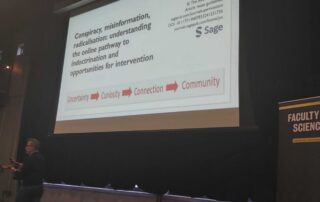 A person stands in a lecture hall pointing at a screen displaying a presentation titled "Conspiracy, misinformation, radicalisation: understanding the online pathway to indoctrination and opportunities for intervention," led by Canada Research Chair Timothy Caulfield. A banner reads "Faculty of Science.