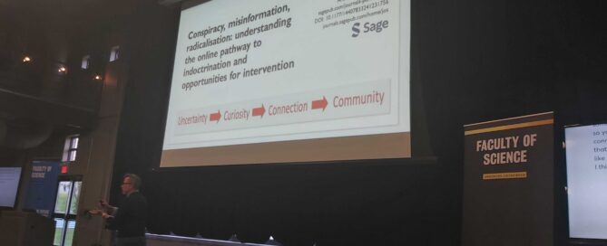 A person stands in a lecture hall pointing at a screen displaying a presentation titled "Conspiracy, misinformation, radicalisation: understanding the online pathway to indoctrination and opportunities for intervention," led by Canada Research Chair Timothy Caulfield. A banner reads "Faculty of Science.