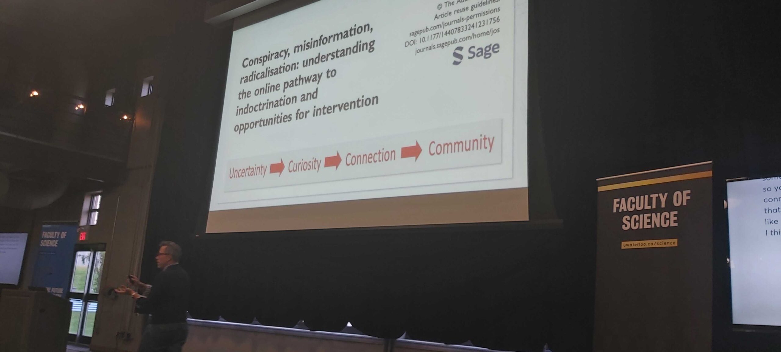 A person stands in a lecture hall pointing at a screen displaying a presentation titled "Conspiracy, misinformation, radicalisation: understanding the online pathway to indoctrination and opportunities for intervention," led by Canada Research Chair Timothy Caulfield. A banner reads "Faculty of Science.