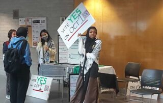 A group of students set up an informational table in a hallway. One holds a sign reading "Vote Yes on Oct 1." Other signs and flyers about divestment are displayed on the table. Some students wear masks and converse, rallying their peers in what appears to be a school or campus setting.