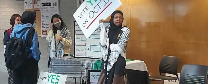 A group of students set up an informational table in a hallway. One holds a sign reading "Vote Yes on Oct 1." Other signs and flyers about divestment are displayed on the table. Some students wear masks and converse, rallying their peers in what appears to be a school or campus setting.