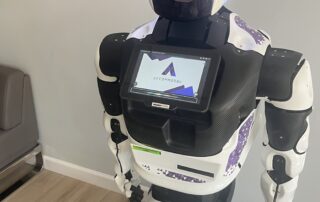 A humanoid robot with a screen on its chest displaying the Accomobbu logo stands on a light wood floor in a room with gray walls. Its digital face features animated eyes and a mouth. A sign behind it reads "ACCOMOBBU Virtual Assistant," highlighting its AI-driven support for students seeking living accommodations.