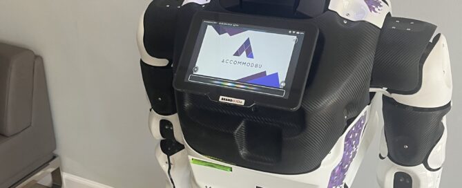 A humanoid robot with a screen on its chest displaying the Accomobbu logo stands on a light wood floor in a room with gray walls. Its digital face features animated eyes and a mouth. A sign behind it reads "ACCOMOBBU Virtual Assistant," highlighting its AI-driven support for students seeking living accommodations.