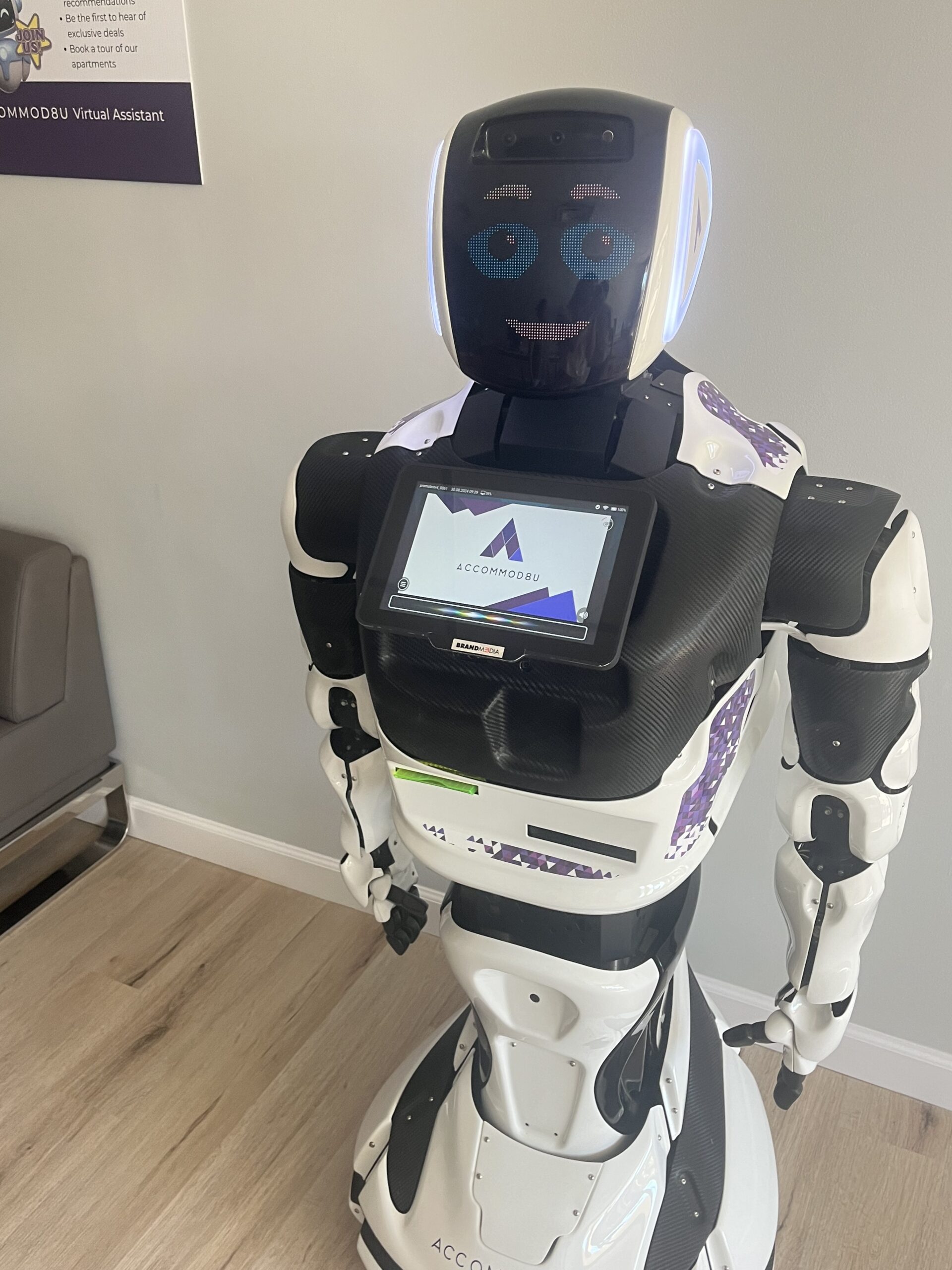 A humanoid robot with a screen on its chest displaying the Accomobbu logo stands on a light wood floor in a room with gray walls. Its digital face features animated eyes and a mouth. A sign behind it reads "ACCOMOBBU Virtual Assistant," highlighting its AI-driven support for students seeking living accommodations.