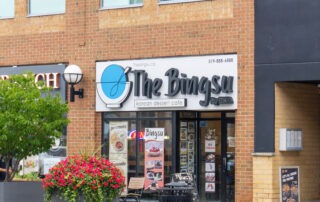 Street view of a brick building featuring "The Bingsu," a Korean dessert café in Waterloo. The storefront displays signs and a menu, with large planters filled with flowers on the sidewalk. The café has a modern design, expertly blending glass and brick elements—a sweet guide to indulgence.