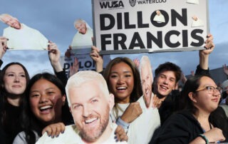 A group of enthusiastic fans are gathered, holding cutouts and posters of a man with short blond hair and a beard. One large sign reads, "Dillon Francis" with smaller text above showing "WUSA" and "University of Waterloo." The fans are smiling and excited, eagerly anticipating the campus concert.