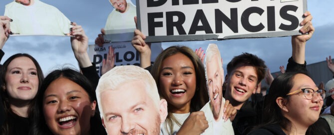 A group of enthusiastic fans are gathered, holding cutouts and posters of a man with short blond hair and a beard. One large sign reads, "Dillon Francis" with smaller text above showing "WUSA" and "University of Waterloo." The fans are smiling and excited, eagerly anticipating the campus concert.