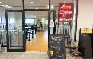 A glass door entrance to a cafe named "Math Coffee & Donuts" with a neon sign welcomes those looking to eat on campus. A chalkboard by the door lists today's soups: Klondike Chili and Vegetable Chili. Inside are tables and chairs, and a single person is visible in the background, facing away.