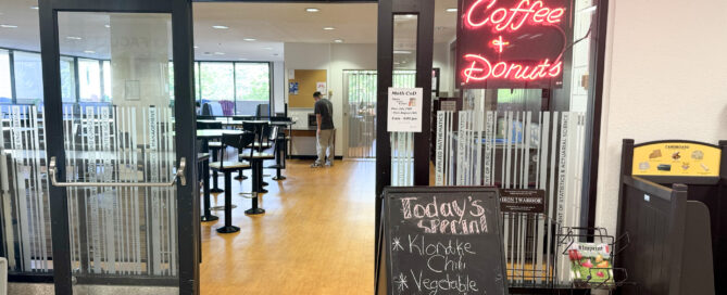 A glass door entrance to a cafe named "Math Coffee & Donuts" with a neon sign welcomes those looking to eat on campus. A chalkboard by the door lists today's soups: Klondike Chili and Vegetable Chili. Inside are tables and chairs, and a single person is visible in the background, facing away.