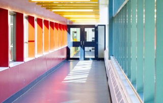 A brightly lit hallway with colorful accents resembles a vibrant campus corridor. Yellow ceiling beams contrast with red and teal walls, while large windows on one side allow sunlight to stream in, casting shadows on the dark floor. At the end, double doors create a gateway to new discoveries.