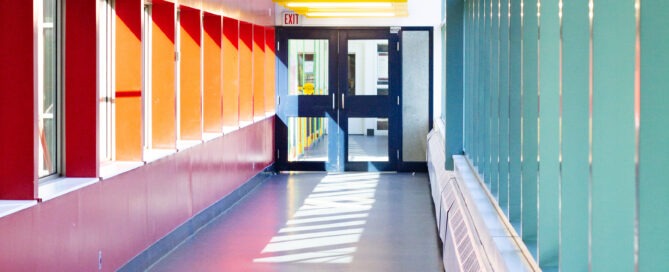 A brightly lit hallway with colorful accents resembles a vibrant campus corridor. Yellow ceiling beams contrast with red and teal walls, while large windows on one side allow sunlight to stream in, casting shadows on the dark floor. At the end, double doors create a gateway to new discoveries.