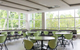 A modern cafeteria with large windows letting in natural light. The room features round tables with light green chairs. The interior design is minimalist, with white walls, a light-colored floor, and a view of greenery outside.