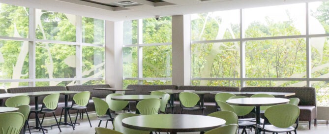 A modern cafeteria with large windows letting in natural light. The room features round tables with light green chairs. The interior design is minimalist, with white walls, a light-colored floor, and a view of greenery outside.