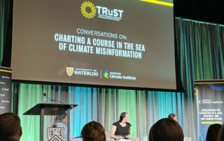 A conference stage with a large screen displaying "Conversations on: Charting a Course in the Sea of Climate Misinformation," hosted by the TRuST Network. A panel discussion like an interactive lecture unfolds with three people seated on stage, while attentive audience members observe from the foreground.