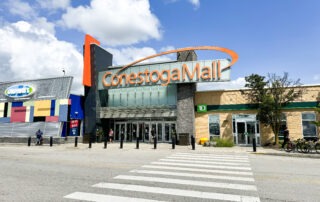 A wide-angle photo of the entrance to Conestoga Mall under a blue sky in the Waterloo region. The mall's orange signage is prominent above the glass doors. Flanking the entrance are a colorful store with "Coppa's" sign and a TD bank branch, offering both shopping excitement and relaxation. A crosswalk is visible in the foreground.