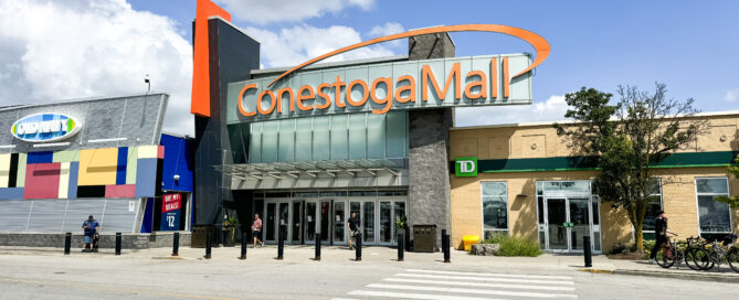 A wide-angle photo of the entrance to Conestoga Mall under a blue sky in the Waterloo region. The mall's orange signage is prominent above the glass doors. Flanking the entrance are a colorful store with "Coppa's" sign and a TD bank branch, offering both shopping excitement and relaxation. A crosswalk is visible in the foreground.