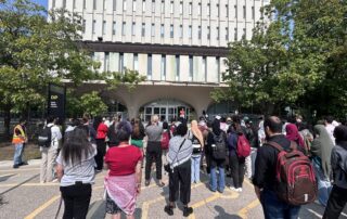 A large group of people stand outside a tall building, gathered around its entrance. Some hold signs while others listen attentively. The gathering, marked by UW Voices for Palestine rallies, appears to be a peaceful assembly or protest. Trees flank both sides of the building, and a sign reads "DP.