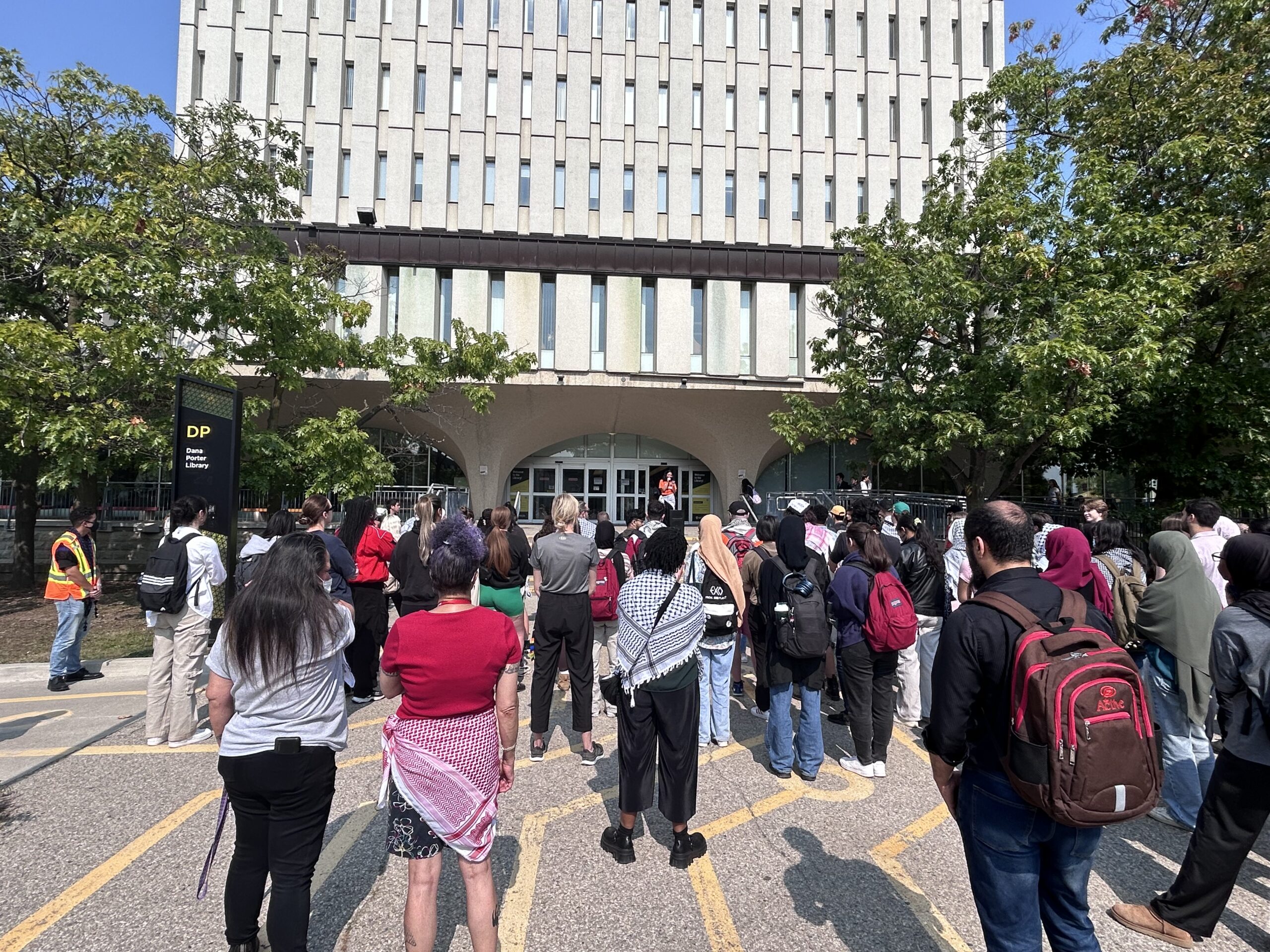 A large group of people stand outside a tall building, gathered around its entrance. Some hold signs while others listen attentively. The gathering, marked by UW Voices for Palestine rallies, appears to be a peaceful assembly or protest. Trees flank both sides of the building, and a sign reads "DP.