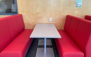 A restaurant booth with red cushioned seats on either side of a rectangular gray table. The booth is set against a light wood-paneled wall with a power outlet, and a framed picture is partially visible on the wall.