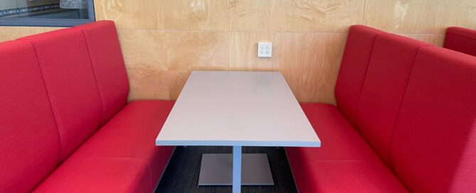 A restaurant booth with red cushioned seats on either side of a rectangular gray table. The booth is set against a light wood-paneled wall with a power outlet, and a framed picture is partially visible on the wall.