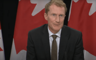 A man in a suit discusses the future of international student permits at a podium, flanked by several Canadian flags, emphasizing changes projected for 2025.