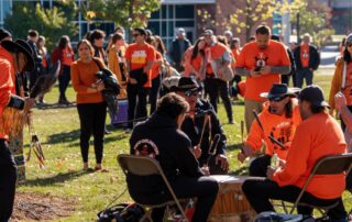 A group of people, many wearing orange shirts, gather outdoors. Several individuals are seated around a drum, playing it, while others stand and watch. The background shows trees and buildings. The scene appears festive with bright sunlight and a joyful atmosphere that hints at reconciliation.