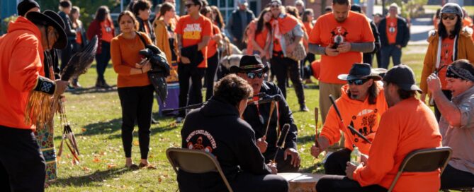 A group of people, many wearing orange shirts, gather outdoors. Several individuals are seated around a drum, playing it, while others stand and watch. The background shows trees and buildings. The scene appears festive with bright sunlight and a joyful atmosphere that hints at reconciliation.