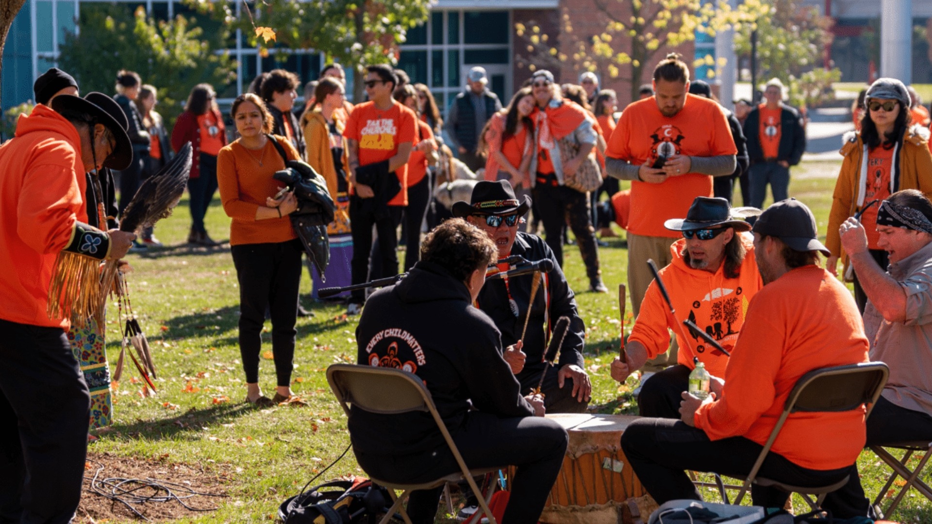 A group of people, many wearing orange shirts, gather outdoors. Several individuals are seated around a drum, playing it, while others stand and watch. The background shows trees and buildings. The scene appears festive with bright sunlight and a joyful atmosphere that hints at reconciliation.