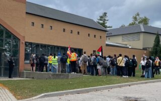 A group of students rally outside a brick building with large windows. Some wear neon vests, and a Palestinian flag waves among them, demanding divestment from UW complicity. Trees and a cloudy sky form the backdrop to their passionate gathering.