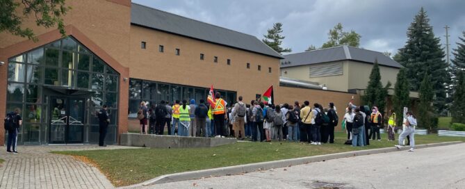 A group of students rally outside a brick building with large windows. Some wear neon vests, and a Palestinian flag waves among them, demanding divestment from UW complicity. Trees and a cloudy sky form the backdrop to their passionate gathering.