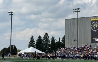 A football game unfolds on a sunny day, watched from the stands behind a railing. The Waterloo Warriors clash with the Laurier Golden Hawks on the field, as spectators fill the bleachers and a tent stands in the background under a cloudy sky during homecoming weekend.