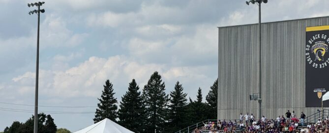 A football game unfolds on a sunny day, watched from the stands behind a railing. The Waterloo Warriors clash with the Laurier Golden Hawks on the field, as spectators fill the bleachers and a tent stands in the background under a cloudy sky during homecoming weekend.