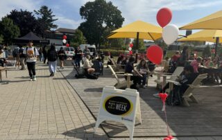 Outdoor event on the arts quad featuring people gathered around tables under yellow umbrellas. Red and white balloons decorate the area. Signage reads "Black and Gold Week." Attendees are seen walking, sitting, eating a free barbecue, and socializing on a sunny day.