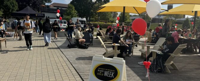 Outdoor event on the arts quad featuring people gathered around tables under yellow umbrellas. Red and white balloons decorate the area. Signage reads "Black and Gold Week." Attendees are seen walking, sitting, eating a free barbecue, and socializing on a sunny day.