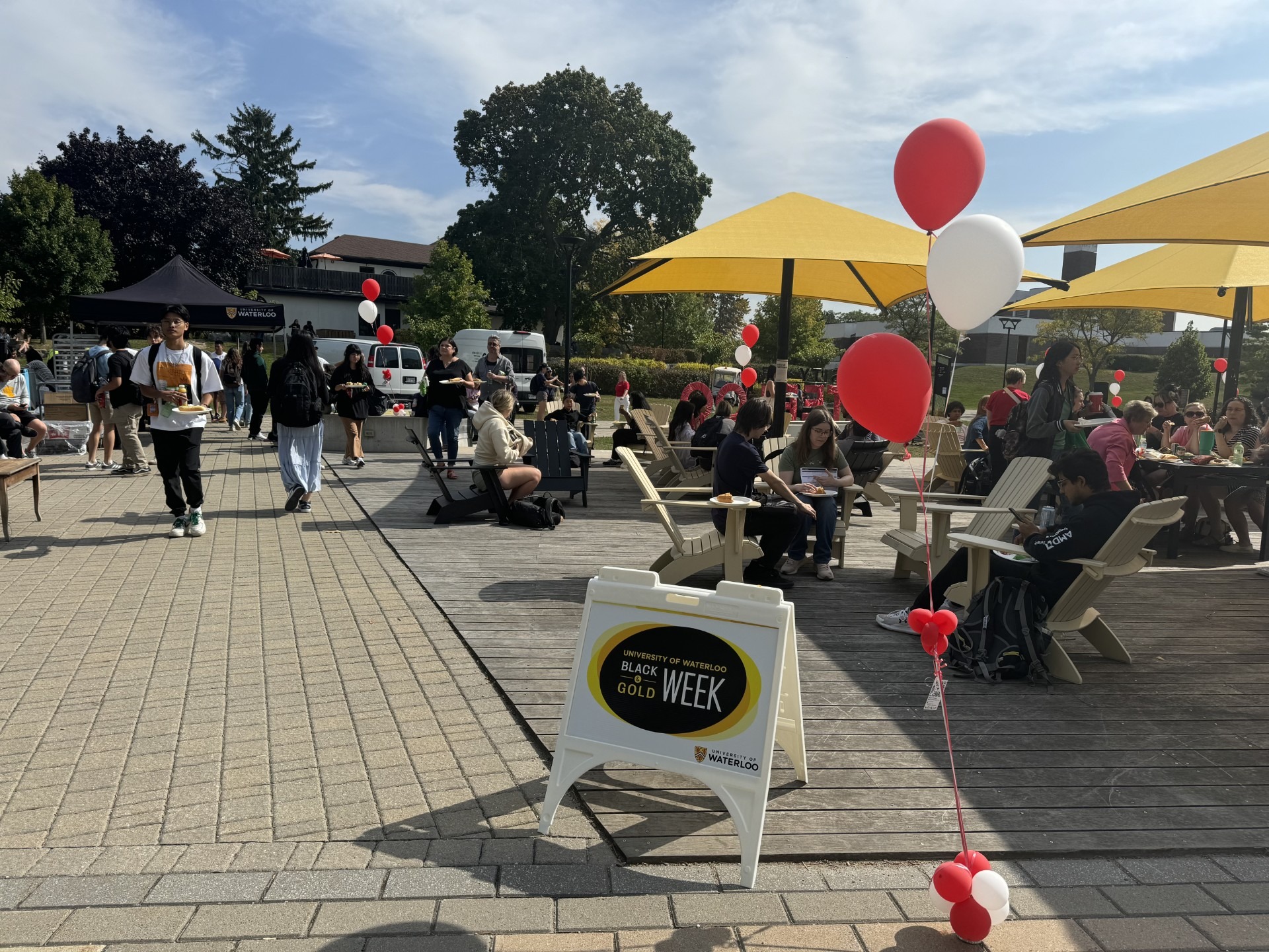 Outdoor event on the arts quad featuring people gathered around tables under yellow umbrellas. Red and white balloons decorate the area. Signage reads "Black and Gold Week." Attendees are seen walking, sitting, eating a free barbecue, and socializing on a sunny day.