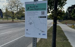 A signpost with a map for the "Route 91 Late Night Loop" stands on the sidewalk, under a partly cloudy sky, with trees lining the street. This successful campaign features a QR code and route details to enhance your late night bus experience.