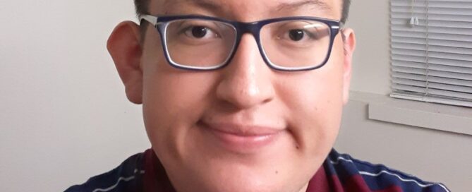 A person wearing glasses and a striped polo shirt, possibly a UW student, is smiling and looking at the camera. The background features a window with blinds and a white wall.