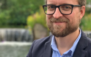 A bearded man with glasses is smiling, standing in front of a blurred background featuring a waterfall and greenery. He is wearing a blue shirt and a dark blazer, resembling the thoughtful demeanor of a biology professor discussing prevention techniques inspired by "The Last of Us.