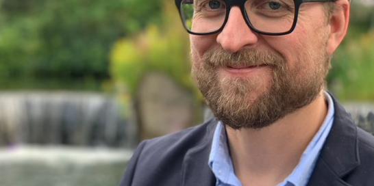 A bearded man with glasses is smiling, standing in front of a blurred background featuring a waterfall and greenery. He is wearing a blue shirt and a dark blazer, resembling the thoughtful demeanor of a biology professor discussing prevention techniques inspired by "The Last of Us.