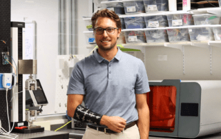 A man wearing glasses and a short-sleeved polo shirt stands in a laboratory. He has a black prosthetic arm, possibly designed during his time as an engineering student. Shelves with containers and various lab equipment surround him, as he researches an innovative lymphedema device.