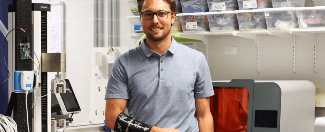 A man wearing glasses and a short-sleeved polo shirt stands in a laboratory. He has a black prosthetic arm, possibly designed during his time as an engineering student. Shelves with containers and various lab equipment surround him, as he researches an innovative lymphedema device.